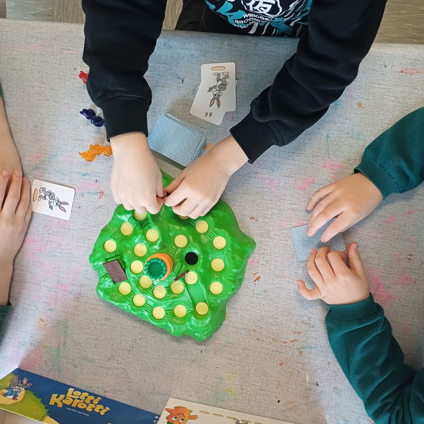 Eine Gruppe von Kindern spielt ein Brettspiel. Das Spiel hat ein grünes Brett mit gelben Kreisen. Die Kinder halten Karten und interagieren mit den Spielsteinen.
