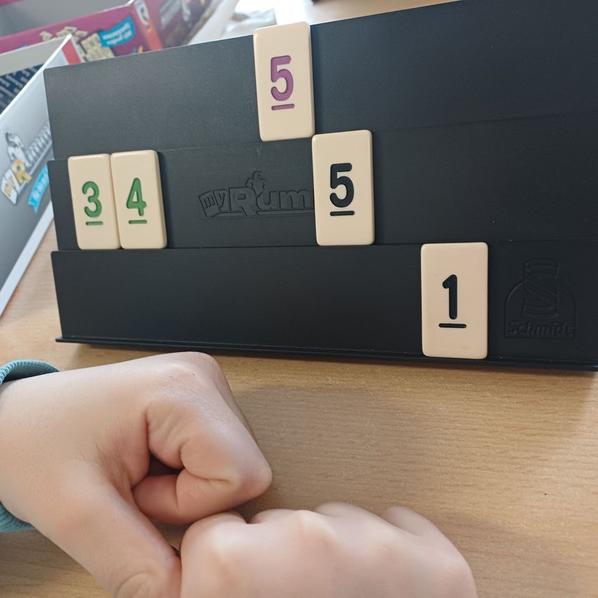 Eine Person spielt ein Brettspiel mit einem schwarzen Brett, auf dem die Zahlen 1, 3, 4 und 5 angezeigt werden. Die Person hält Würfel und scheint dabei zu sein, sie zu werfen.