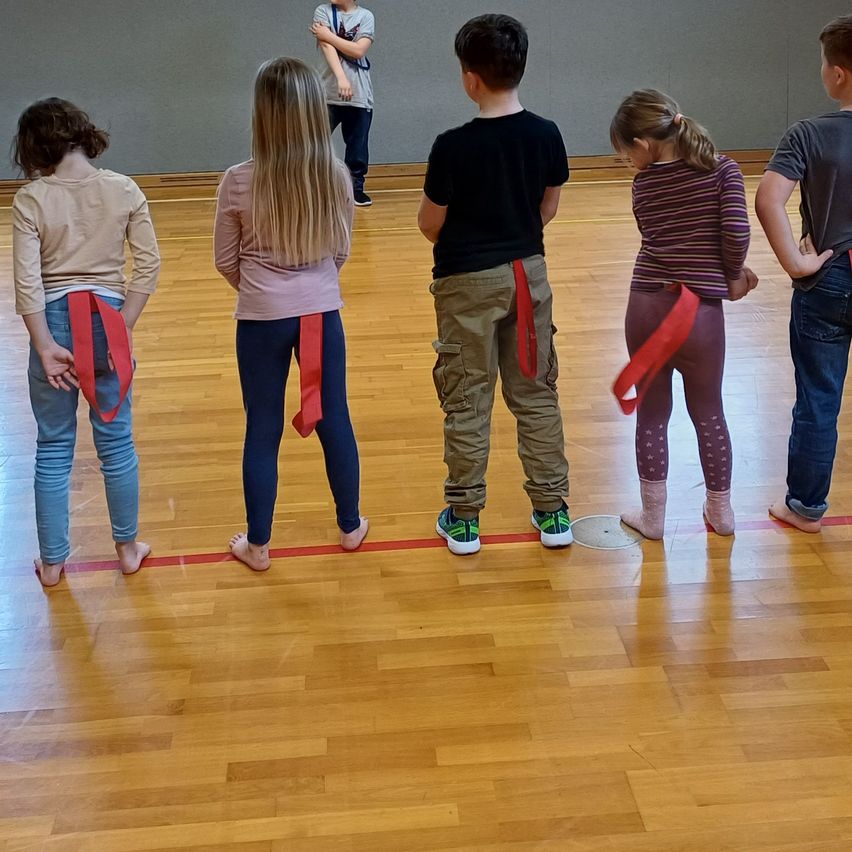 Fünf Kinder mit roten Bändern um die Taille stehen in einer Reihe auf einem Holzboden, ein sechstes Kind im Hintergrund.