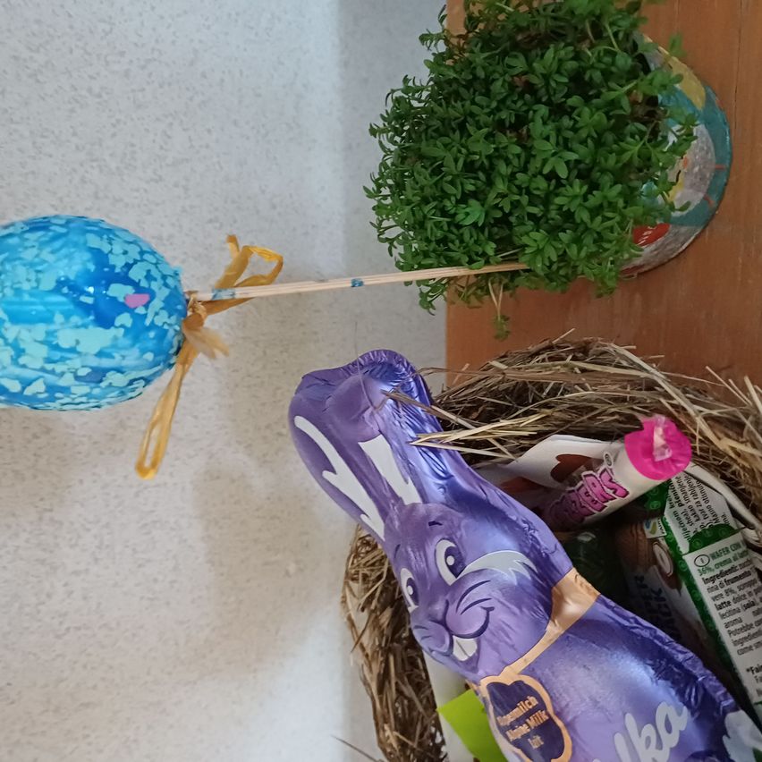 Ein dekoratives Osterei mit einer Schleife befindet sich neben einer Pflanze. Ein Korb mit einer Osterhasen-Schokolade ist in der Nähe.