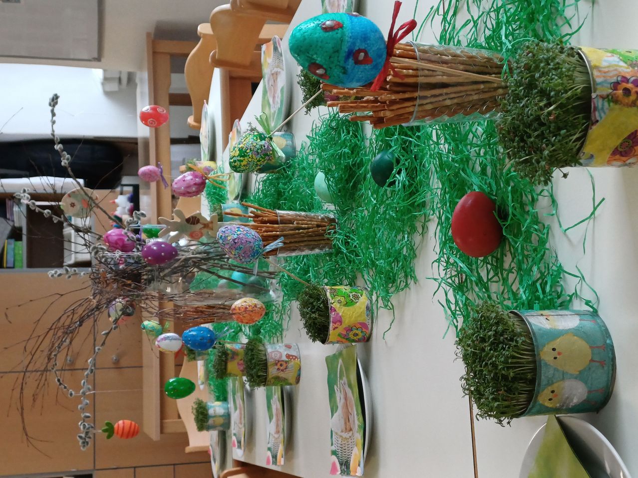 Eine weiße Wand, die mit grünem Gras, verschiedenen Ostereiern und Papierbasteleien geschmückt ist. Der Tisch ist mit einem blauen Ei und einer roten Schleife auf Krackern eingerichtet.