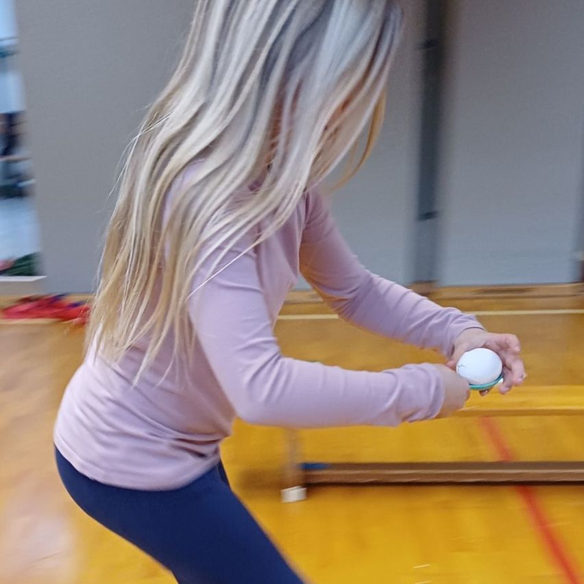 Eine Frau in einem rosa Shirt und blauen Hosen spielt Tischtennis und schwingt ihren Schläger. Der Ball ist in der Luft, und die Szene spielt sich in einer Turnhalle ab.