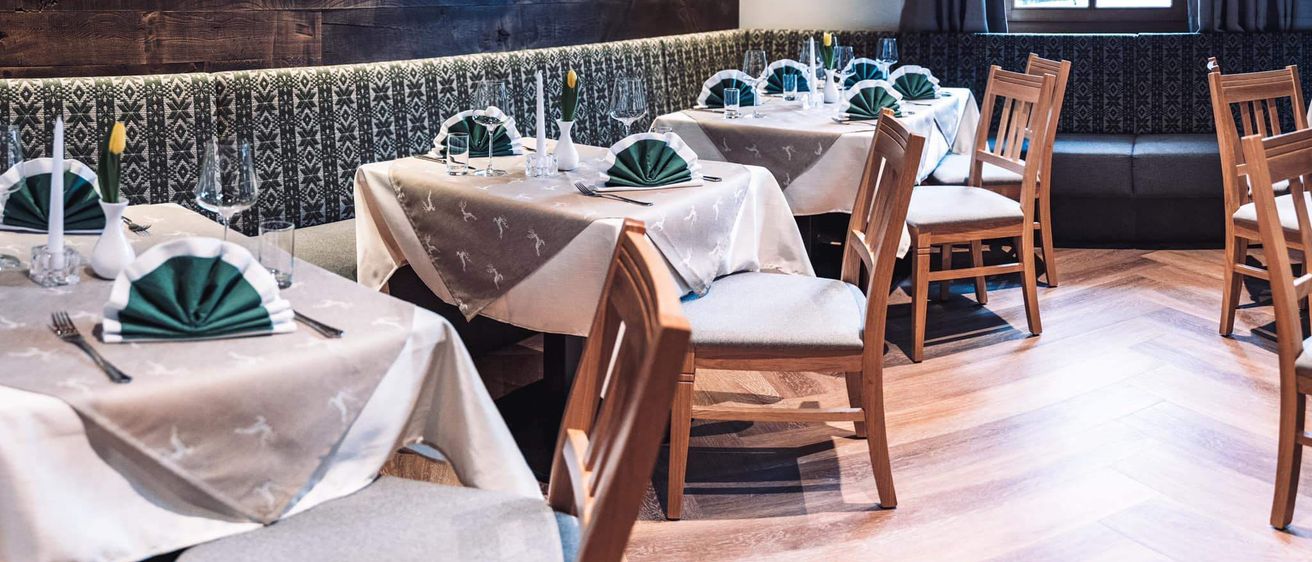 Ein gemütliches Restaurant mit Holzwänden und -böden, das zwei Tische für das Essen bietet. Jeder Tisch hat eine weiße Tischdecke, Weingläser und Servietten. Ein Tisch hat eine Vase mit Blumen. Eine Lampe hängt von der Decke.