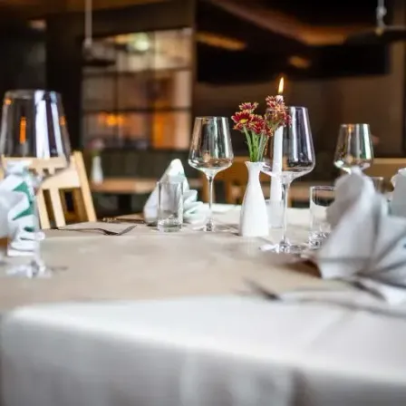 Ein Tisch mit weißer Tischdecke, Weingläsern und Blumen in einer weißen Vase. Eine Kerze ist angezündet.