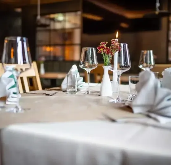 Ein Tisch mit weißer Tischdecke, Weingläsern und Blumen in einer weißen Vase. Eine Kerze ist angezündet.