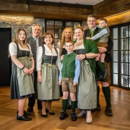 Eine Familie steht in einem Raum in traditioneller deutscher Kleidung, mit einem Holzboden und einer Ziegelmauer hinter ihnen. Sie scheinen für ein Foto zu posieren.