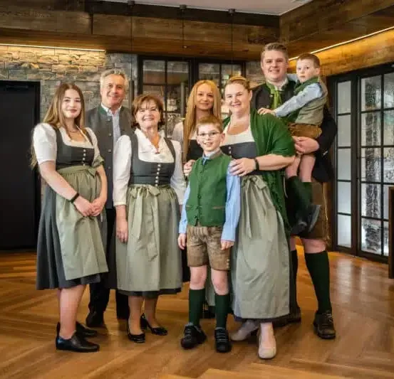 Eine Familie steht in einem Raum in traditioneller deutscher Kleidung, mit einem Holzboden und einer Ziegelmauer hinter ihnen. Sie scheinen für ein Foto zu posieren.