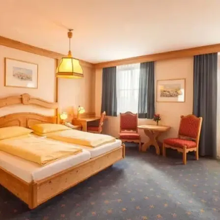 Ein Hotelzimmer mit Holzmöbeln. Ein Doppelbett mit gelben Kissen, ein Schreibtisch mit Stuhl und ein Tisch mit Blumen. Zwei gerahmte Bilder hängen an der Wand und zwei Fenster mit Vorhängen.