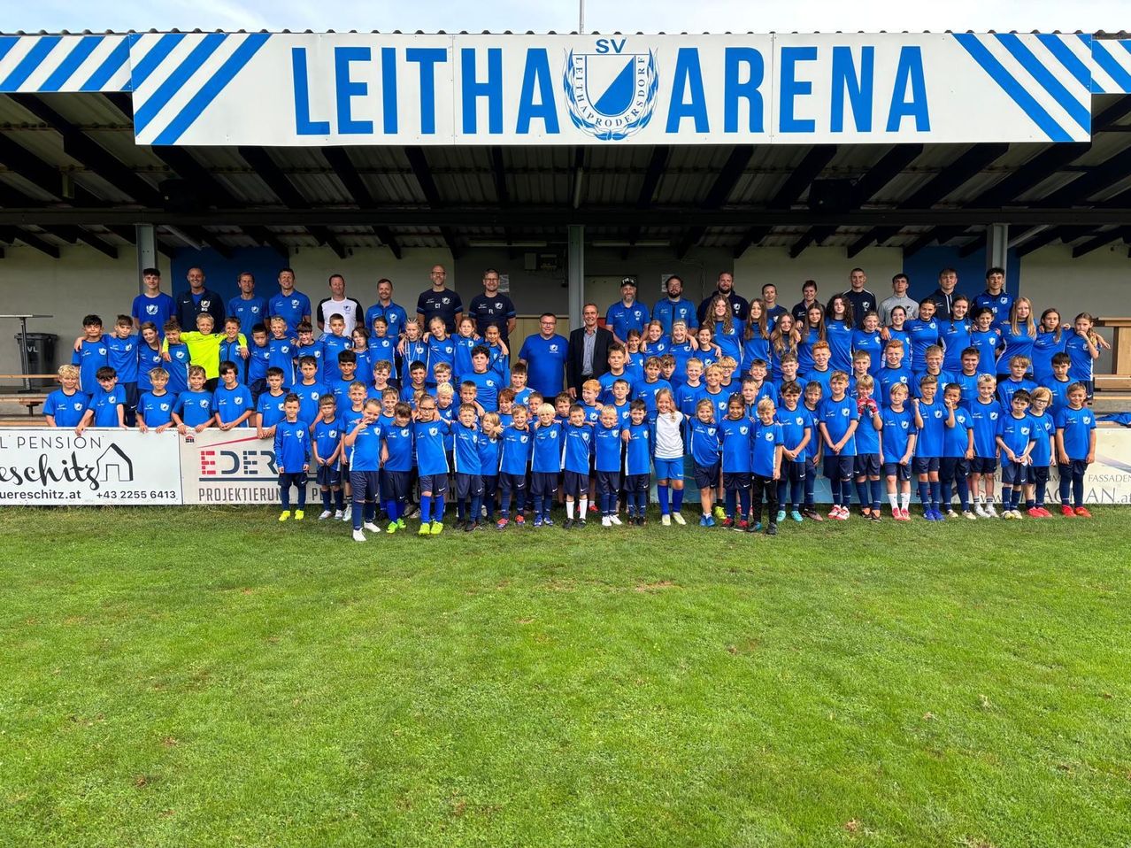 Eine Fußballmannschaft posiert für ein Foto vor einem Feld, mit einem Schild, das 'Leithau Arena' darüber steht. Sie tragen blaue Uniformen, und ein Banner mit dem Wort 'Eder' ist hinter ihnen.
