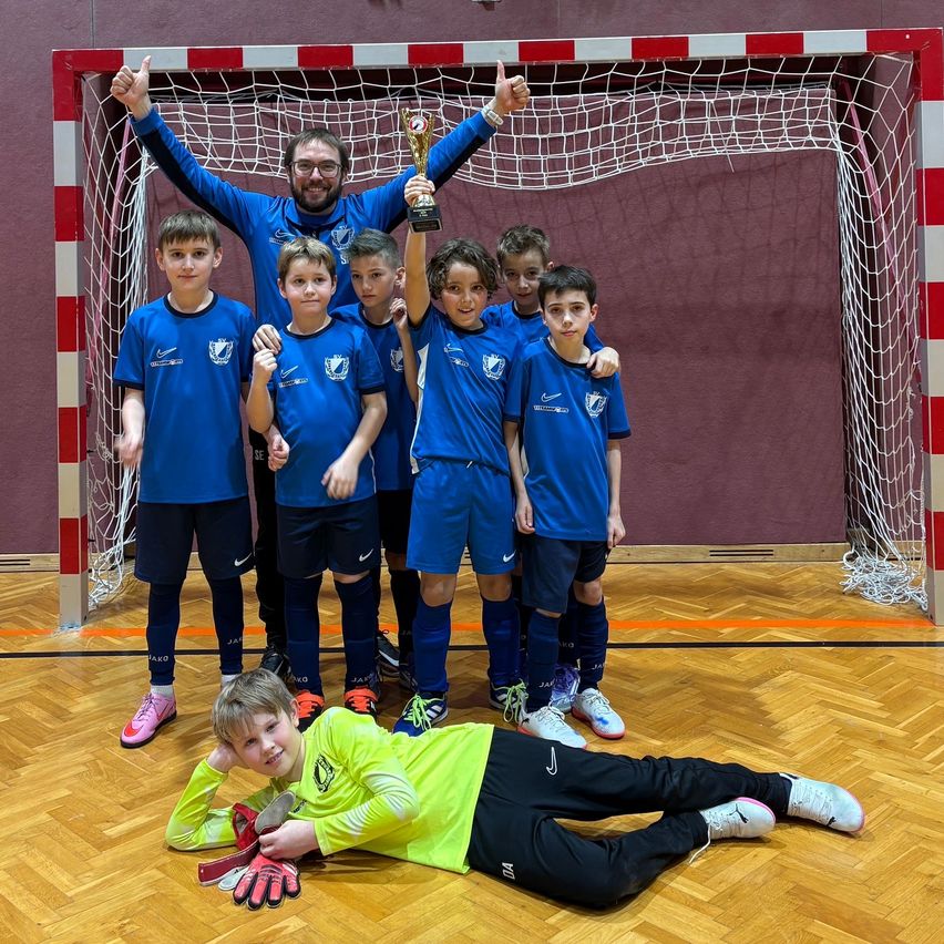 Eine Gruppe junger Fußballspieler und ein Trainer feiern mit einer Trophäe vor einem Tor.