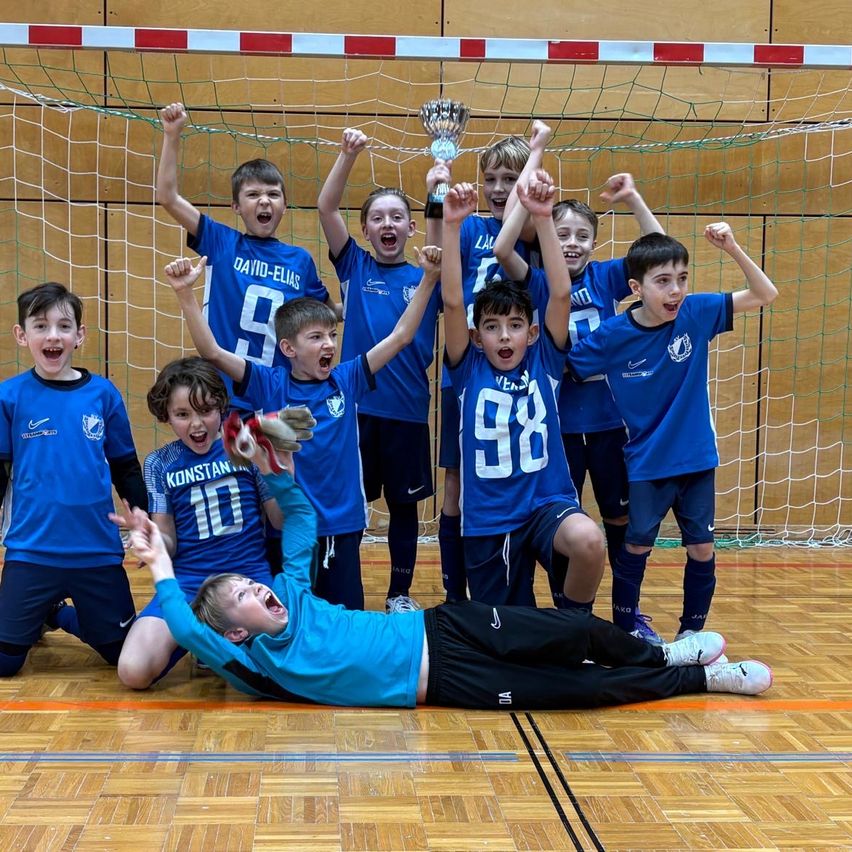 Eine Gruppe junger Jungen in blauen Trikots feiert einen Sieg, wobei ein Junge mit einem Pokal auf dem Boden liegt. Sie stehen hinter einem Netz mit roten und weißen Rändern, und der Junge in der Mitte hat Handschuhe an.