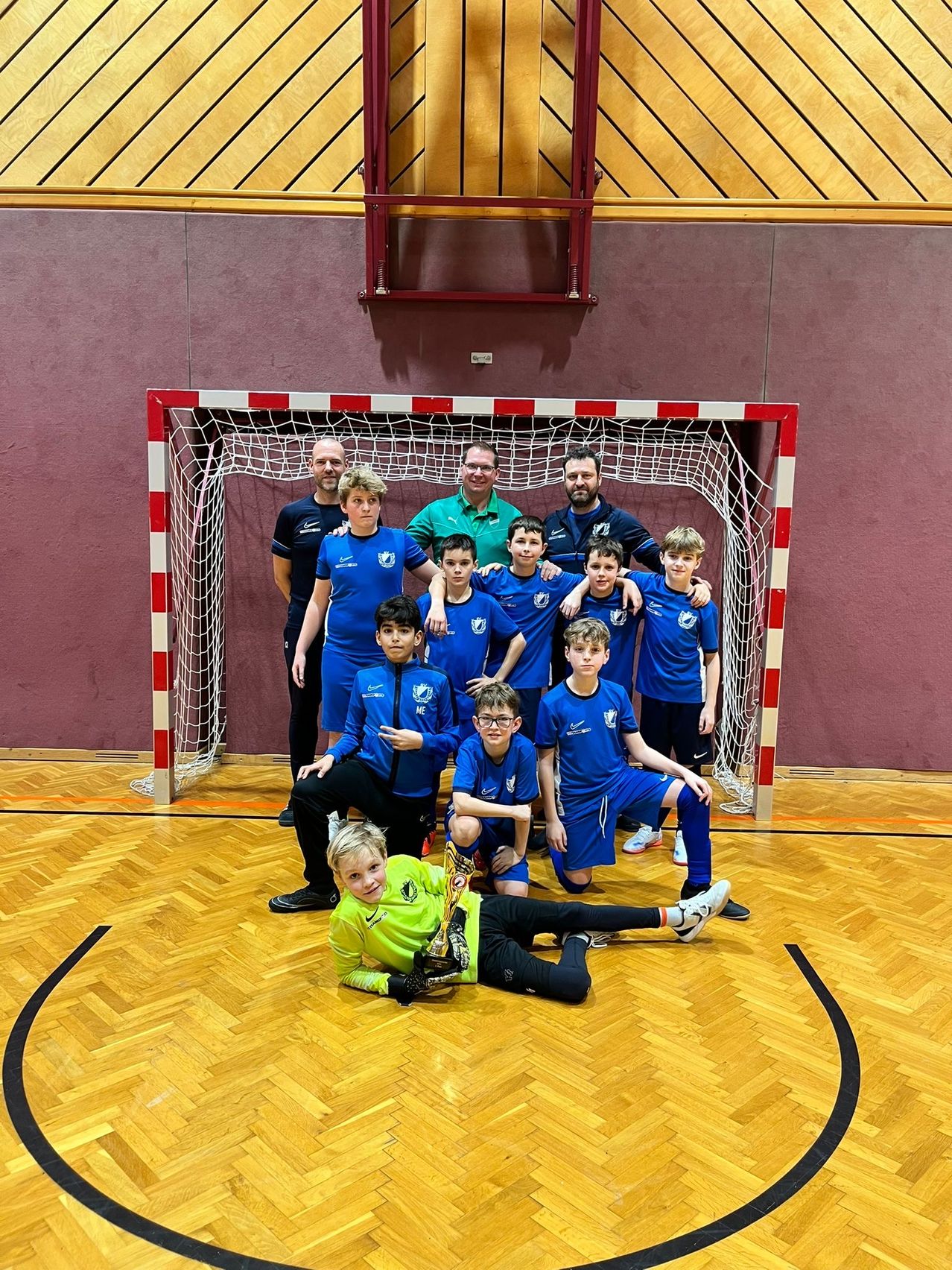 Eine Gruppe junger Fußballspieler in blauen Trikots posiert mit ihren Trainern vor einem Tor für ein Foto. Die Spieler halten einen Pokal.