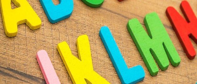 Das Bild zeigt eine Sammlung bunter hölzerner Buchstaben, die auf einer Holzoberfläche angeordnet sind. Die Buchstaben sind in verschiedenen Farben und Größen und einige sind kopfüber. Der Hintergrund ist eine Holzstruktur.