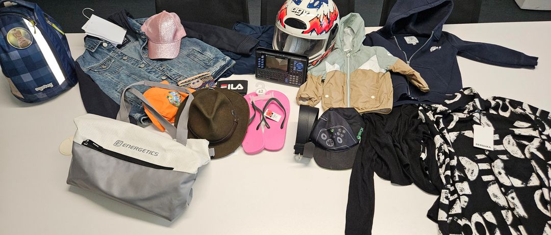 Auf einem Tisch sind ein Helm, eine Jeansjacke, eine rosa Kappe, eine Kapuzenjacke, eine graue Tasche und andere Gegenstände zu sehen.