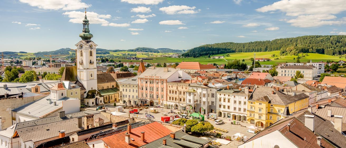 Luftaufnahme einer europäischen Stadt mit einem hohen Glockenturm, verschiedenen Gebäuden und üppigen grünen Hügeln in der Ferne.