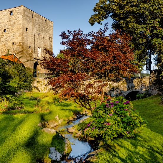 Ein großes Steingebäude mit einem gewölbten Eingang, umgeben von üppigem Grün und einem kleinen Bach.