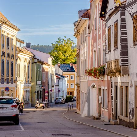 Eine europäische Straße mit geparkten Autos, Gebäuden und Bäumen. Die Gebäude haben verschiedene Designs und Farben, mit einem Baum in der Mitte. Ein Roller und Fahrräder sind an der Straßenseite geparkt.