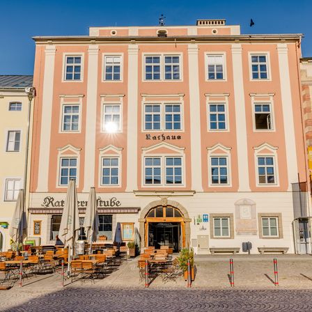 Ein mehrstöckiges Gebäude mit einem Restaurant und Sitzgelegenheiten im Freien. Das Gebäude trägt den Namen 'Rathaus'. Draußen stehen Sonnenschirme und Tische.