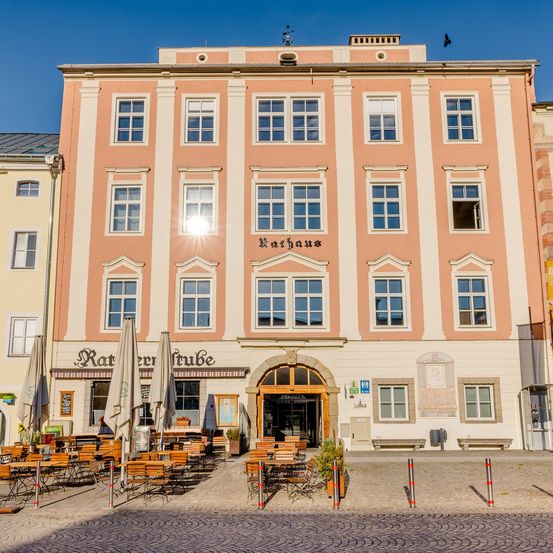 Ein mehrstöckiges Gebäude mit einem Restaurant und Sitzgelegenheiten im Freien. Das Gebäude trägt den Namen 'Rathaus'. Draußen stehen Sonnenschirme und Tische.