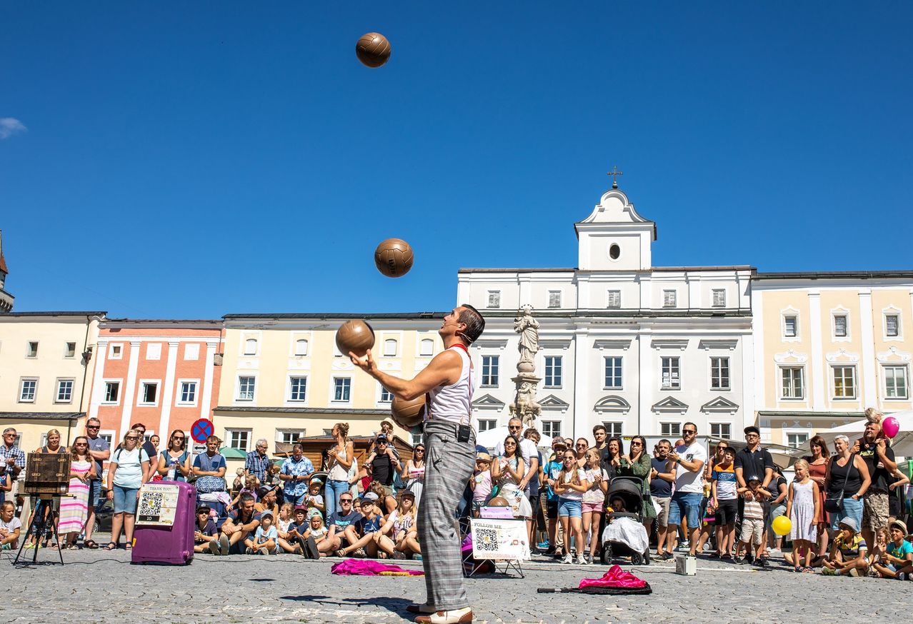 Ein Mann jongliert mit drei Basketballbällen in der Mitte eines Stadtplatzes, während eine große Menschenmenge zusieht. Die Gebäude hinter ihm sind in verschiedenen Farben gehalten, wobei ein markantes weißes Gebäude im Hintergrund steht.