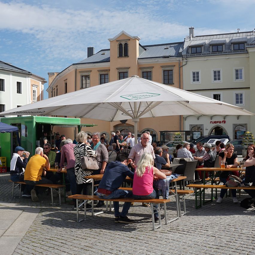 Eine Gruppe von Menschen sitzt unter einem großen Schirm an Tischen in einer Außenumgebung, mit Gebäuden im Hintergrund.