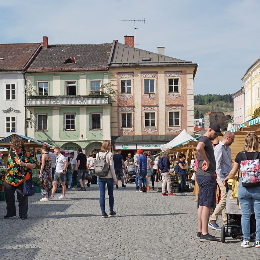 Ein Stadtplatz mit Menschen, Geschäften und Gebäuden. Die Straße ist mit Kopfsteinpflaster gepflastert.