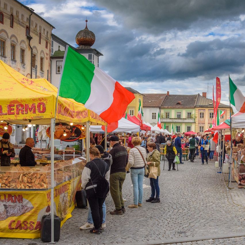 Ein Straßenmarkt mit verschiedenen Essensständen, einer mit einer großen italienischen Flagge. Mehrere Menschen durchstöbern die Stände unter einem bewölkten Himmel.
