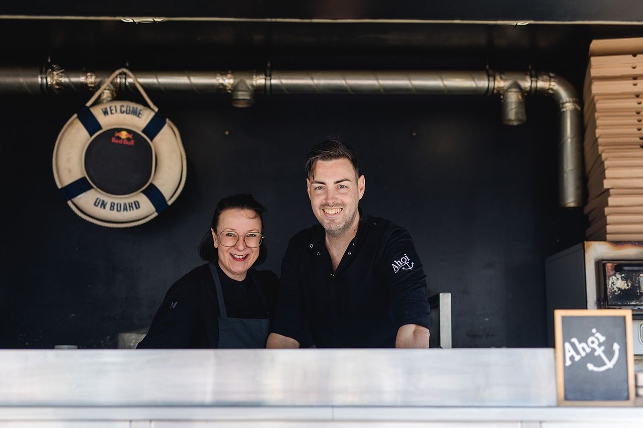 Zwei Personen stehen lächelnd hinter einem Tresen. Ein Rettungsring hängt an der Wand hinter ihnen. Der Mann trägt ein schwarzes Shirt mit Logo.
