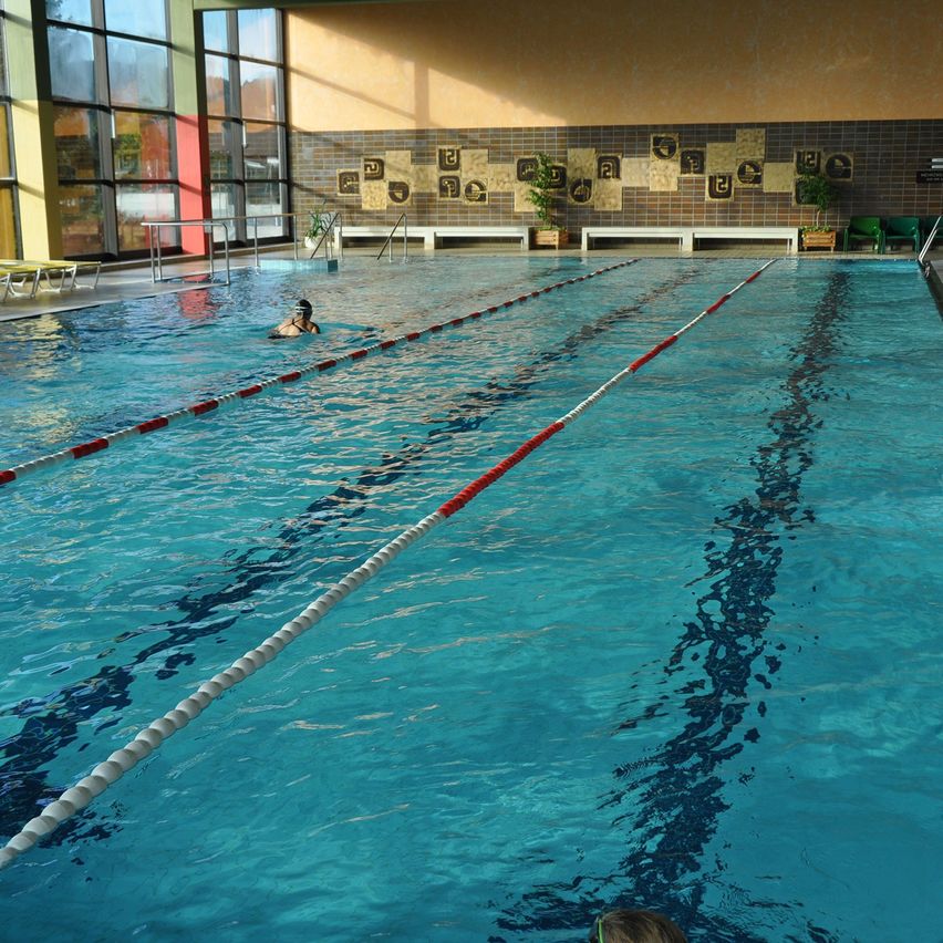 Ein Hallenschwimmbad mit durch weiße und rote Seile markierten Bahnen. Eine Person schwimmt im Becken, während eine andere Person von der Seite zuschaut. Das Becken hat Glaswände und eine Wand mit gefliesten Designs.
