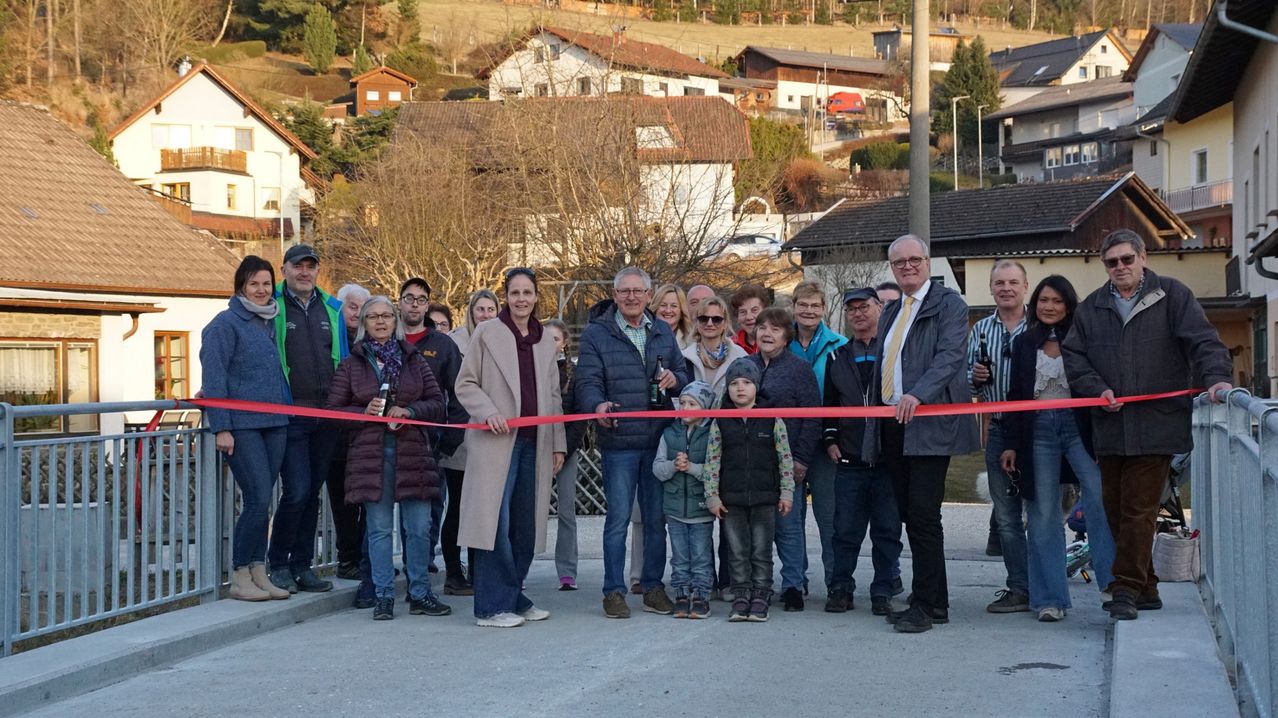 Eine Gruppe von Menschen, einschließlich Kinder, steht hinter einem roten Band, das geschnitten wird, in einem Außenbereich. Die Veranstaltung markiert wahrscheinlich eine gemeinschaftliche Feier oder Eröffnung.