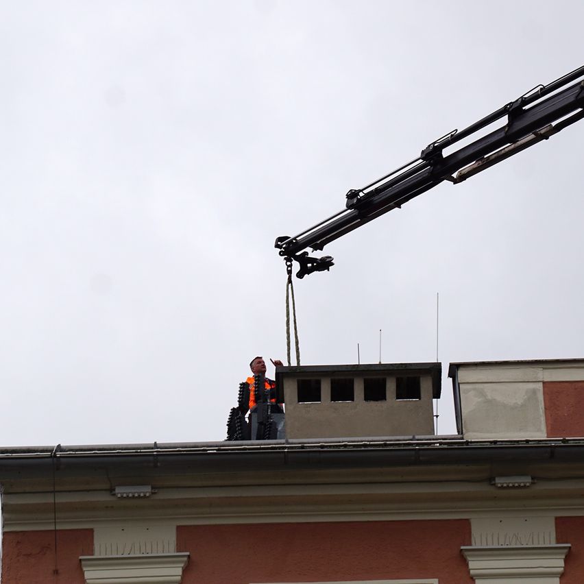 Ein Mann in einer orangefarbenen Warnweste steht auf einem Dach und wird von einem Kran angehoben. Das Gebäude hat einen Schornstein und der Himmel ist bedeckt.