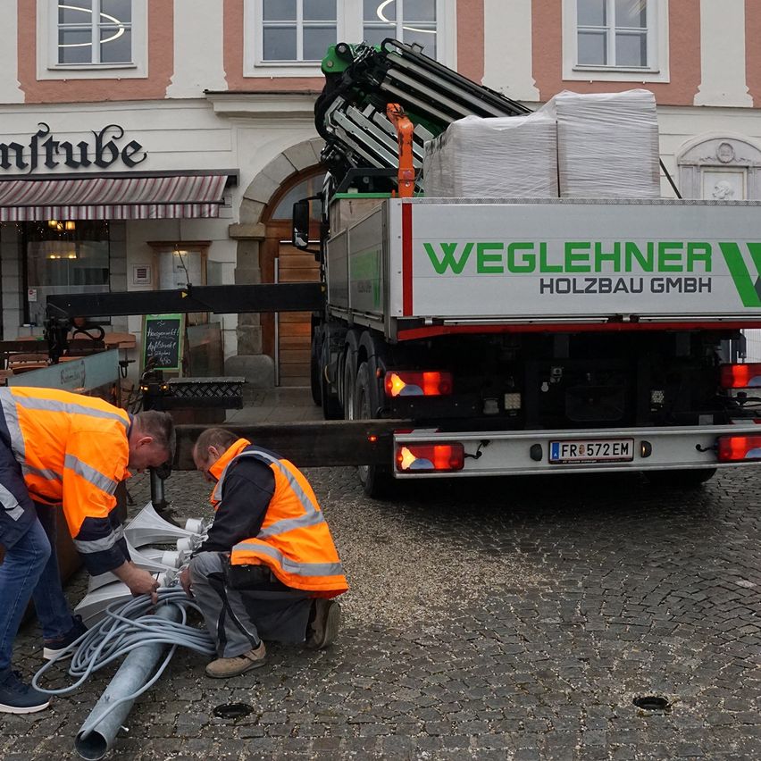 Zwei Arbeiter in orangen Westen entladen Waren von einem Lkw mit dem Namen Weglehner Holzbau GmbH. Der Lkw ist vor einem Gebäude mit einem gestreiften Vordach geparkt.