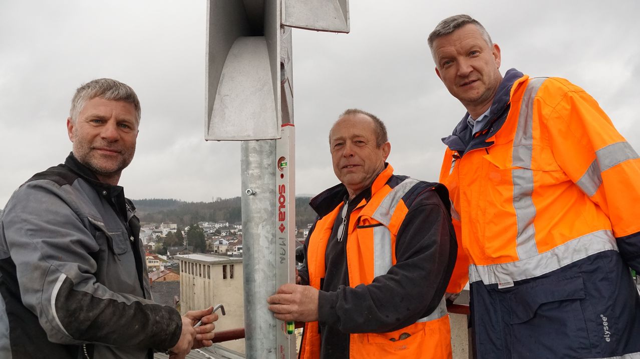 Drei Männer in orangefarbenen Westen stehen auf einem Dach mit einem Metallpfahl. Ein Mann hält ein Werkzeug. Im Hintergrund sind Gebäude und Hügel zu sehen.