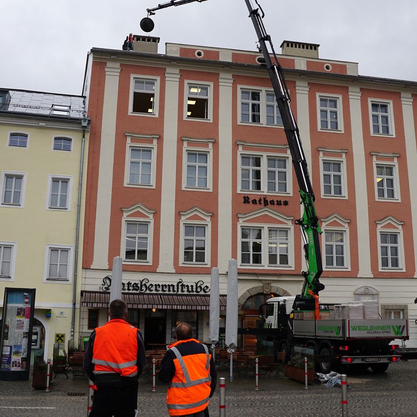 Zwei Arbeiter in Warnwesten stehen in der Nähe eines Krans, der ein schweres Objekt hebt. Das Gebäude hat viele Fenster und das Wort Rathaus ist sichtbar.