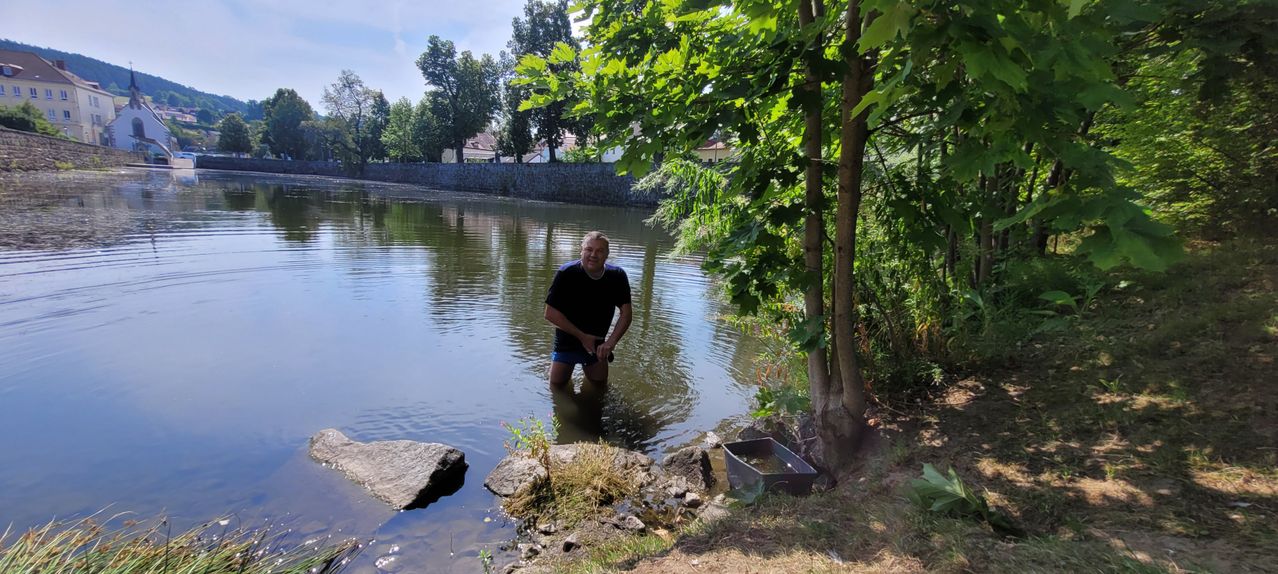 A man stands in a river, smiling, with a small boat nearby and a tree trunk on the right.