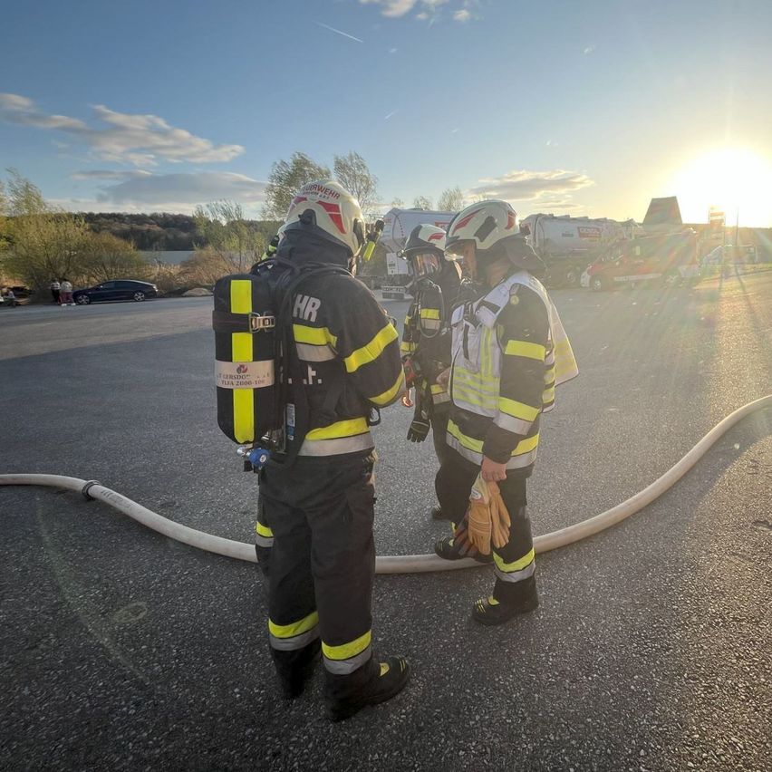Drei Feuerwehrleute in voller Ausrüstung stehen auf einem Parkplatz, ein Feuerwehrschlauch liegt auf dem Boden und die untergehende Sonne ist im Hintergrund zu sehen.