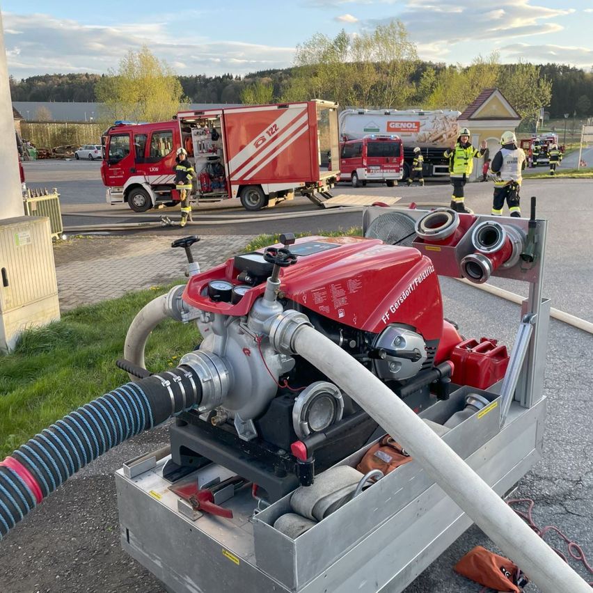 Feuerwehrleute stehen neben einem Feuerwehrwagen und einem Anhänger, beide auf einem gepflasterten Bereich geparkt. Der Wagen hat seine Tür offen, und eine große rote Pumpe ist davor positioniert. Der Himmel ist bewölkt.