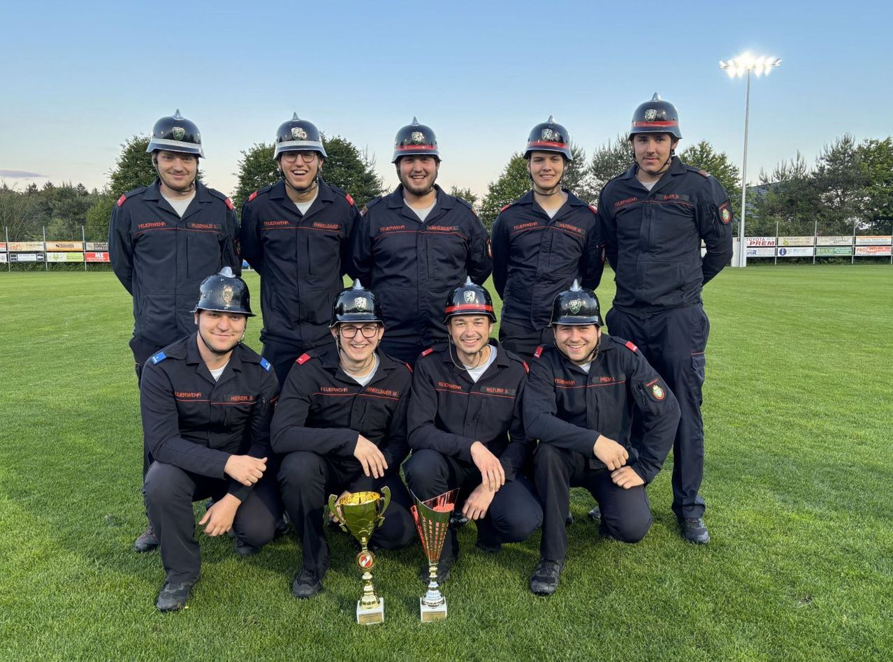 Eine Gruppe Feuerwehrleute in Uniform posiert für ein Foto auf einem Feld mit zwei Trophäen vor ihnen. Sie lächeln und scheinen einen Sieg zu feiern.