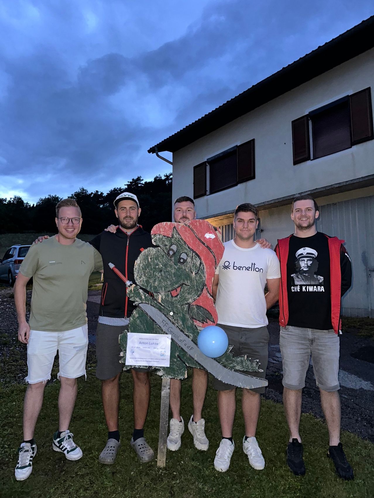 Fünf Männer stehen vor einem Haus, einer hält ein Schild, ein anderer eine Kettensäge. Sie posieren mit einer grünen Figur. Zwei tragen Jacken, zwei tragen Shorts. Der Himmel ist bewölkt.
