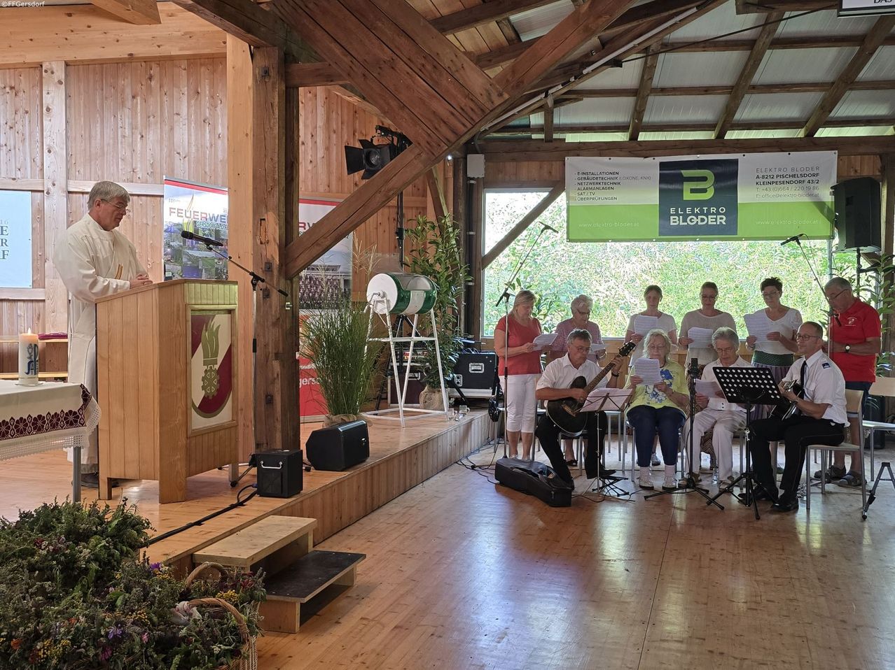 Eine Gruppe von Menschen tritt in einer rustikalen Umgebung auf, ein Mann spielt Gitarre und singt, während andere Papiere halten und zuhören. Ein Banner und eine Topfpflanze sind auf der Bühne.