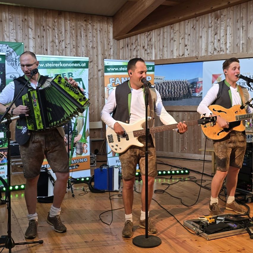 Drei Männer treten auf der Bühne auf. Der Mann auf der linken Seite spielt Akkordeon, der Mann in der Mitte spielt Gitarre und der Mann auf der rechten Seite singt. Sie tragen alle Shorts. Hinter ihnen ist eine Wand mit Werbung.