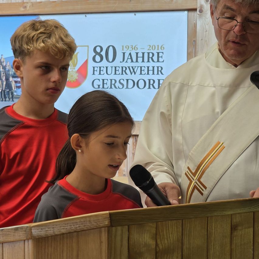 Ein junges Mädchen spricht in ein Mikrofon an einem Podium, während ein Priester und ein Junge zusehen, mit einem Banner im Hintergrund, das 80 Jahre der Feuerwehr Gersdhore feiert.