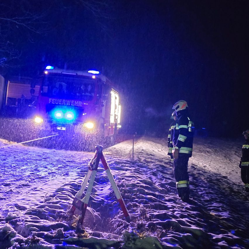 Ein Feuerwehrauto steht bei Nacht auf einer verschneiten Straße, mit Feuerwehrleuten in der Nähe. Das Auto hat blaue Lichter und das Wort 'Feuerwehr' an der Seite.