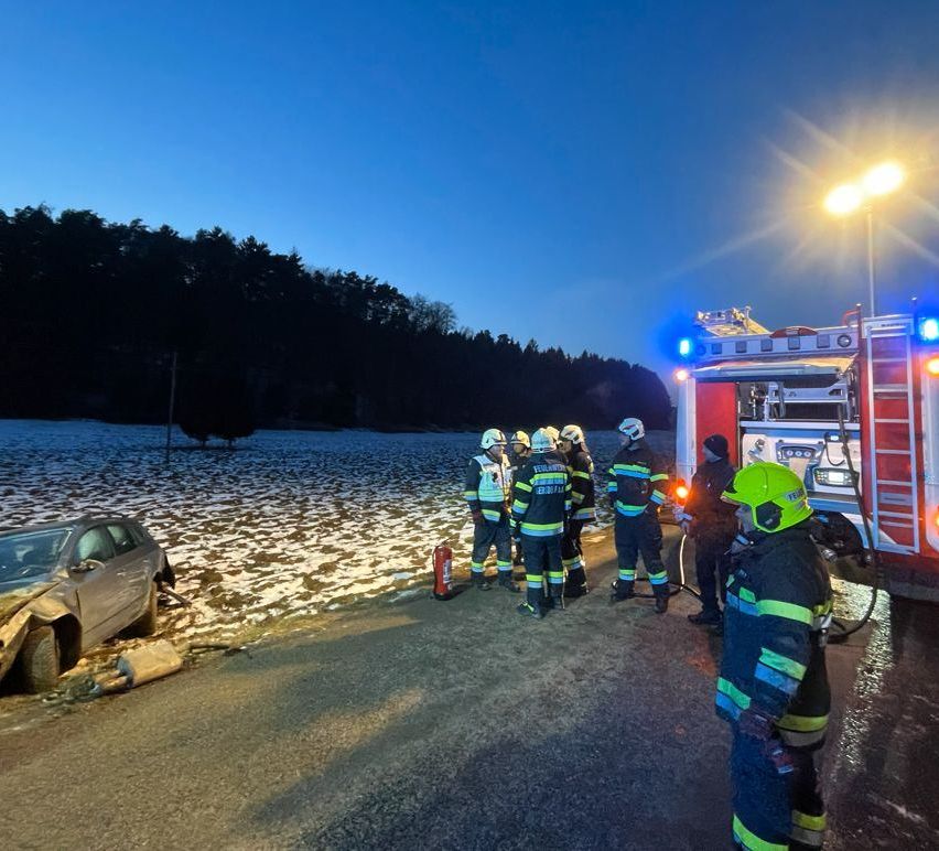 Firefighters are gathered around a wrecked car on a snowy road at dusk. Trees and a bright light are in the background.