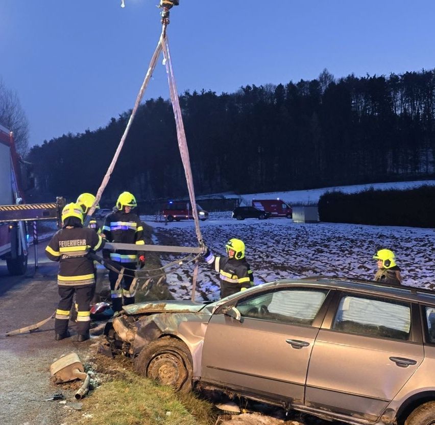 Firefighters work at a crash site, a car is lifted by a crane, with emergency vehicles and a snowy landscape in the background.