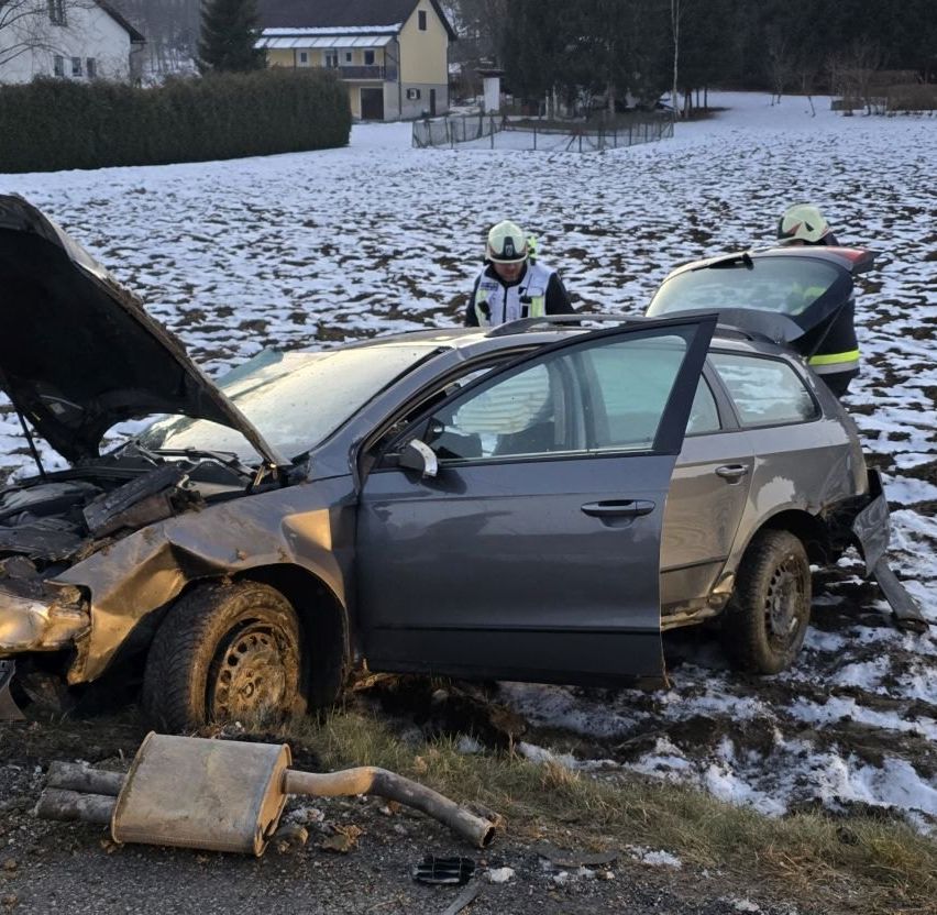 A damaged car with its hood open lies on a snowy road, with two people in safety gear nearby.