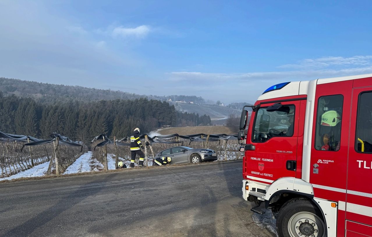 Ein Feuerwehrfahrzeug ist am Straßenrand geparkt, wo zwei Feuerwehrleute sich um ein silbernes Auto kümmern, das in einen Zaun in der Nähe eines Weinbergs gecrasht ist.