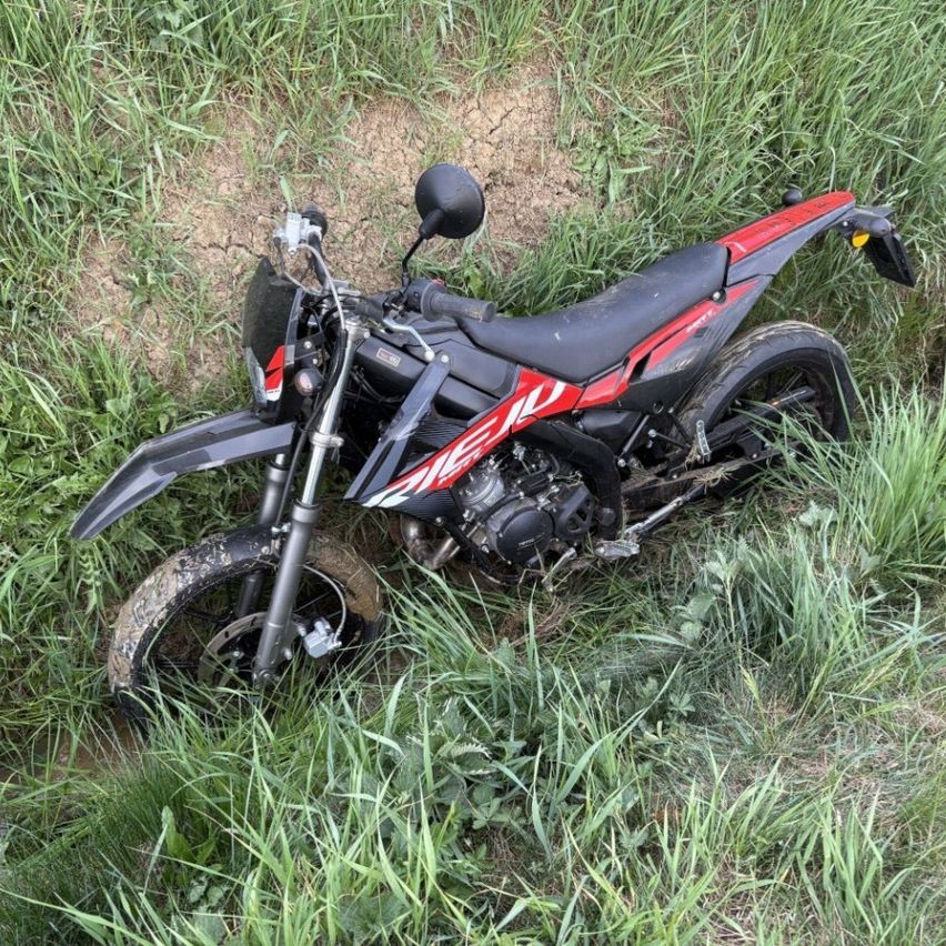 Ein rotes und schwarzes Motorrad liegt auf der Seite in einem Grasfeld. Das Vorderrad ist abmontiert, und das Motorrad ist mit Schmutz und Gras bedeckt.