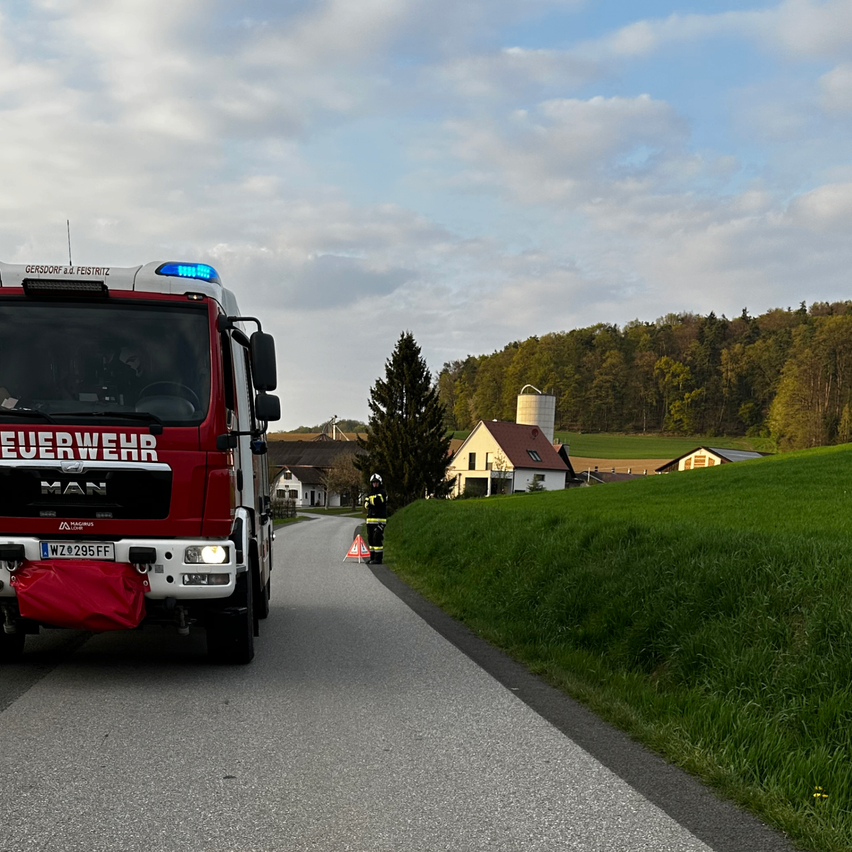 Ein roter Feuerwehrwagen fährt mit einem Warnkegel an der Seite auf einer Straße, eine Person in einem Helm steht daneben. Es gibt Häuser und eine Scheune am Grashang.