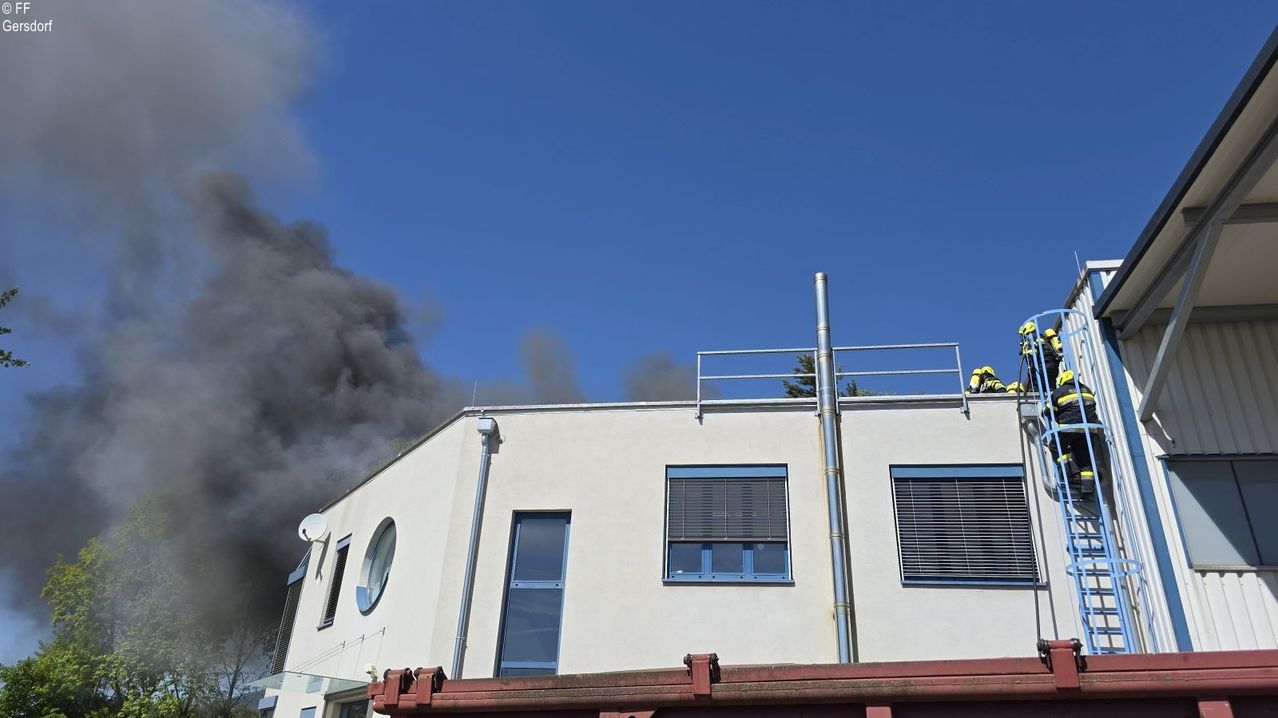 Ein Gebäude mit Rauch, der aus der Spitze quillt, Feuerwehrleute auf dem Dach, ein klarer blauer Himmel im Hintergrund.